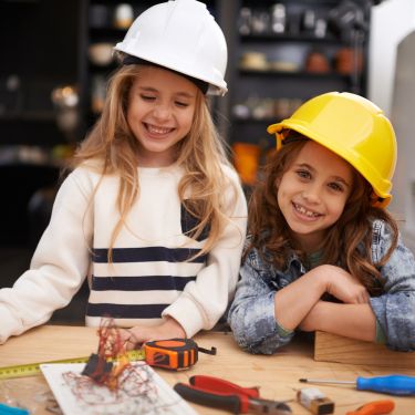 Zwei Mädchen mit gelben Bauhelmen hantieren spielerisch mit Holz und Werkzeug an einer Werkbank.